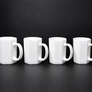 May include: Four white, geometric-shaped mugs with curved handles are arranged in a row against a black background. The mugs have a modern, minimalist design, suitable for coffee, tea, or other beverages.