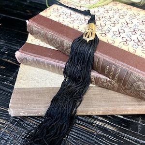 May include: A black tassel with a gold metal top featuring the number "70" is placed on top of a stack of three vintage books. The books have brown leather covers with intricate floral patterns.