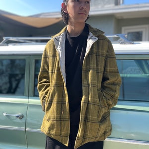May include: A young person wearing a black and yellow plaid jacket with a black baseball cap. The jacket has a zipper closure and a white lining. The person is standing in front of a vintage car.