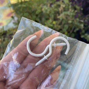 Może przedstawiać: Dwa białe plastikowe haczyki z wkrętami. Haczyki znajdują się w przezroczystym plastikowym woreczku.