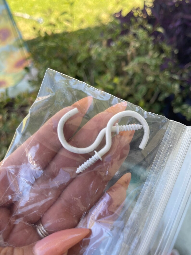 Może przedstawiać: Dwa białe plastikowe haczyki z wkrętami. Haczyki znajdują się w przezroczystym plastikowym woreczku.