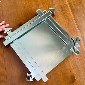 May include: A square mirror with a decorative frame made of mirrored strips. The strips are arranged in a geometric pattern with arrows pointing in different directions.