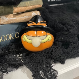 May include: A ceramic figurine of the orange cartoon cat Garfield wearing a black witch hat. The character has a round orange face, narrow eyes, a small white nose, and a wide grin. The figurine is smooth and glossy, featuring the iconic feline features.