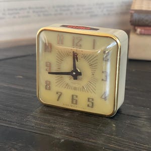May include: A vintage, cream-colored alarm clock with gold accents and a black face. The clock has a square shape and a clear glass front. The clock face has black numbers and hands, and a gold ring around the edge. The clock is made in the USA.