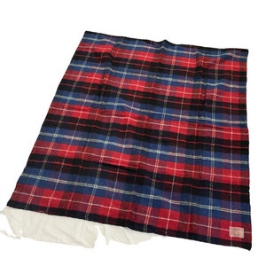 May include: A red, black, and blue plaid blanket. The blanket has a classic plaid pattern with thick stripes of red, black, and blue. The blanket is made of a soft material and is folded. A small tag is visible on the bottom right corner.