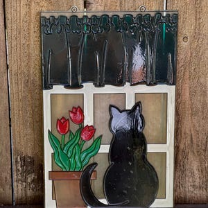 May include: A vintage-style wall hanging features a black cat sitting by a window.  The window has a dark green curtain and a potted plant with three red tulips. The artwork is done in a stained-glass style with dark outlines and bold colors. The piece is framed with a clear plastic border and has two small holes at the top for hanging. The overall theme is whimsical and nostalgic.