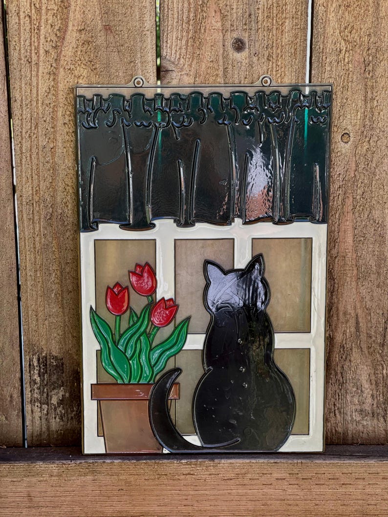 May include: A vintage-style wall hanging features a black cat sitting by a window.  The window has a dark green curtain and a potted plant with three red tulips. The artwork is done in a stained-glass style with dark outlines and bold colors. The piece is framed with a clear plastic border and has two small holes at the top for hanging. The overall theme is whimsical and nostalgic.