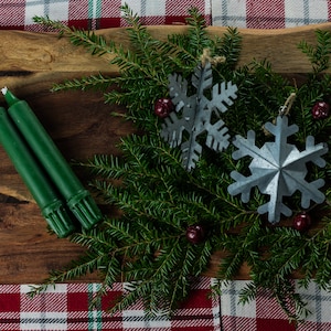 Puede incluir: Dos velas verdes sobre una tabla de cortar de madera con una rama de ramas de hoja perenne y dos adornos de copo de nieve plateado. Los adornos están decorados con bayas rojas.