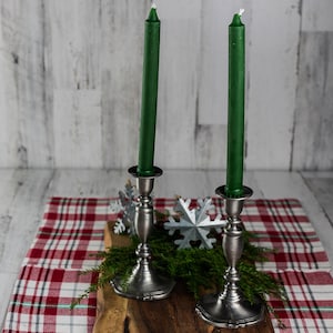 Puede incluir: Dos velas verdes en candelabros de plata sobre una tabla de madera con un mantel a cuadros y decoraciones de copo de nieve plateadas.