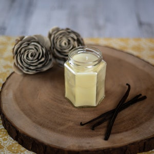 Puede incluir: Una vela en un tarro de vidrio transparente con cera amarilla en el interior, sentada sobre una rodaja de madera con vainas de vainilla. La vela est&aacute; rodeada de dos formas de flores de madera decorativas.