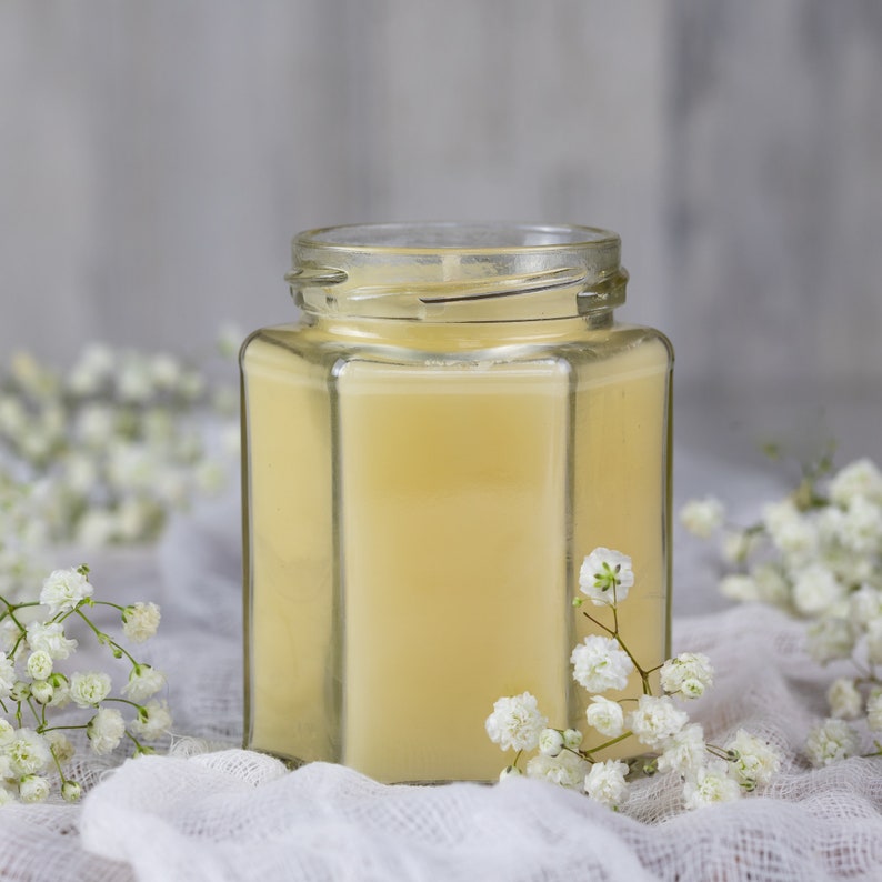 K&ouml;nnte beinhalten: Eine Kerze in einem durchsichtigen Glasgef&auml;&szlig; mit gelber Wachsf&uuml;llung. Die Kerze steht auf einer wei&szlig;en Stoffoberfl&auml;che mit wei&szlig;en Blumen.