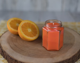 May include: A hexagonal glass jar candle with a bright orange scent. The candle is sitting on a wooden cutting board with two orange halves.