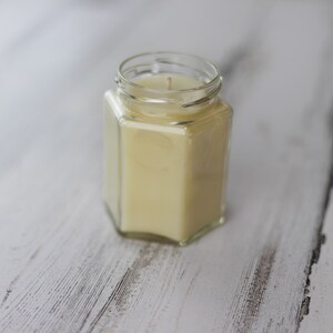 Puede incluir: Una vela en un tarro de vidrio transparente con aroma de cera amarilla. La vela est&aacute; sobre una superficie de madera blanca.