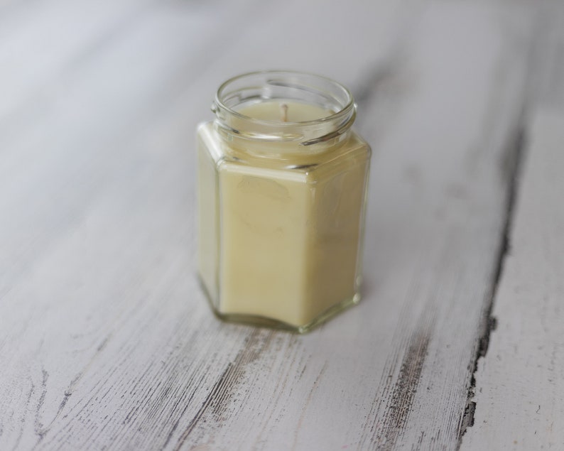 Puede incluir: Una vela en un tarro de vidrio transparente con aroma de cera amarilla. La vela est&aacute; sobre una superficie de madera blanca.