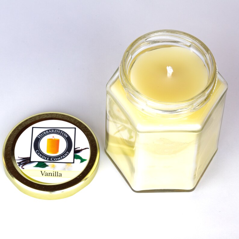 Puede incluir: Una vela en un tarro de vidrio hexagonal con una tapa dorada. La tapa tiene una etiqueta que dice "Hubbardston Candle Company" y "Vanilla". La vela es de color amarillo claro y tiene una sola mecha.