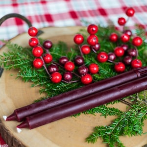 May include: Two burgundy taper candles on a wooden tray with sprigs of evergreen and red berries.