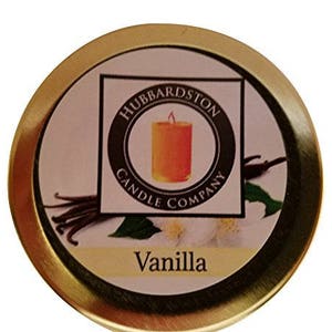 Puede incluir: Una lata dorada con una etiqueta que dice "Hubbardston Candle Company" y "Vanilla". La etiqueta presenta un dibujo de una vaina de vainilla y una vela.