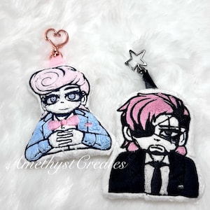 Może przedstawiać: Dwa haftowane breloczki do kluczy z postaciami. Jeden przedstawia postać z różowymi włosami, niebieską kurtką i różową muszką. Drugi ma różowe włosy, czarny garnitur i przepaskę na oku. Oba mają metalowe kółka na klucze.