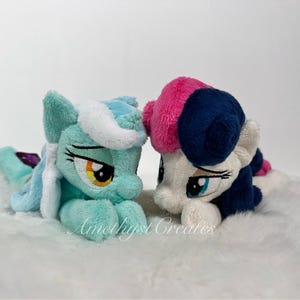 May include: Two plush toys of My Little Pony characters. The pony on the left is light blue with white fur and yellow eyes. The pony on the right is white with pink, blue, and white fur and blue eyes.