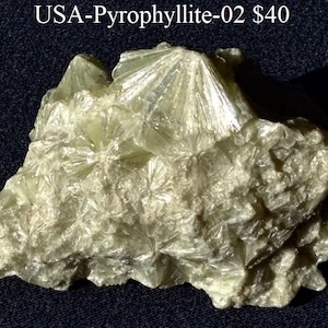 Puede incluir: Un espécimen mineral de pirofilita verde claro con una estructura cristalina. La superficie tiene una apariencia texturizada con patrones de estrella. El texto "USA-Pyrophyllite-02" es visible en la parte superior.