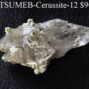 May include: A translucent, crystalline mineral specimen labeled TSUMEB-Cerussite-12. The cluster features elongated, sharp, clear-to-white prismatic crystals with visible striations and small patches of light green mineral deposits embedded within.