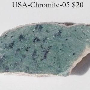 May include: A piece of chromite stone with a light green hue and dark green speckles. The stone has a rough, uneven edge and the text "USA-Chromite-05" is visible at the top.