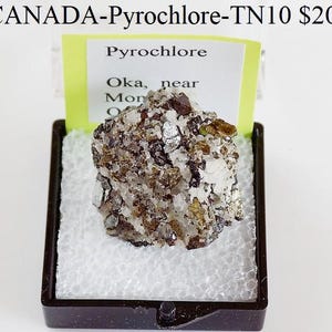 May include: A Pyrochlore mineral specimen from Oka, near Montreal, Canada, presented in a black display case. The mineral features a mix of brown, white, and metallic silver crystals, with a yellow label.