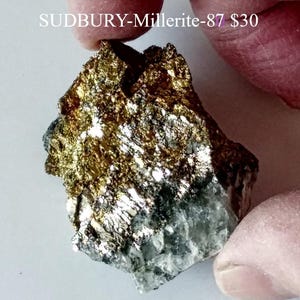 May include: A mineral specimen of Sudbury Millerite-87. The rock has a metallic luster with a mix of gold, silver, and dark gray colors. The surface is rough and textured, with visible crystalline structures. The text "SUDBURY-Millerite-87" is visible.