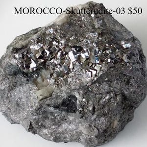 May include: A rough, grey Skutterudite mineral specimen from Morocco. The surface is covered with shiny, metallic crystals. The text "MOROCCO-Skutterudite-03" is visible at the top of the image.