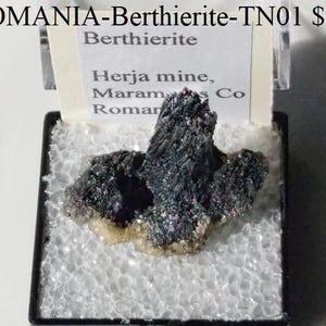 Puede incluir: Un espécimen mineral de Berthierita de Rumanía, exhibido en una caja transparente. El mineral tiene un brillo oscuro y metálico con una estructura cristalina. El espécimen está sobre una base blanca texturizada, con una etiqueta visible.