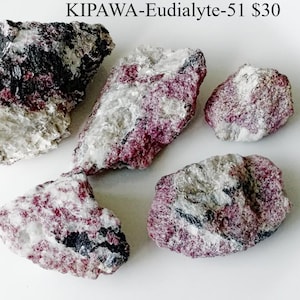 Op de afbeelding: Vijf ruwe eudialiet mineraalmonsters. De stenen hebben een mix van kleuren, waaronder diep roze, wit en zwart. De tekst "KIPAWA-Eudialyte-51" is zichtbaar bovenaan de afbeelding.