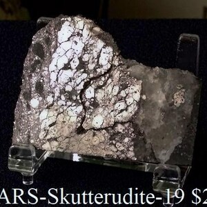 May include: A grey and white rock specimen with a cracked, textured surface, presented on a clear acrylic stand. The rock's surface is marked with a network of fissures. The text "BiARS-Skutterudite-19" is visible at the bottom.
