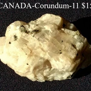 Op de afbeelding: Een close-up van een lichtgekleurd korund exemplaar uit Canada. Het mineraal heeft een gelaagde, kristallijne structuur met kleine zwarte insluitsels. De achtergrond is een donker, getextureerd oppervlak.