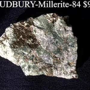 May include: A mineral specimen labeled "SUDBURY-Millerite-84". The rock has a rough, irregular shape with a mix of gray, green, and gold colors. The surface appears textured, with visible crystalline structures. The overall appearance suggests a natural, unpolished stone.