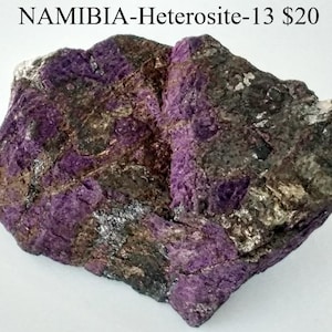 Op de afbeelding: Een ruwe, onregelmatig gevormde steen met een levendige paarse kleur, afgewisseld met grijze, bruine en zilveren delen. De tekst "NAMIBIA-Heterosite-13" is zichtbaar. Dit natuurlijke mineraal heeft een getextureerd oppervlak.