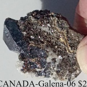 May include: A dark gray galena crystal with a metallic luster, featuring a prominent, sharp crystal face. The mineral specimen has a rough, textured matrix with hints of white and brown.