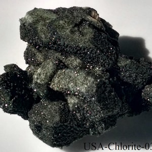 Puede incluir: Un espécimen mineral oscuro y texturizado con una superficie rugosa e irregular. La piedra es principalmente de color gris oscuro con toques de verde e inclusiones brillantes. El texto "USA-Chlorite-03" es visible en la parte inferior.