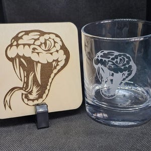 May include: A wooden coaster featuring a laser-engraved image of a snake's head with fangs. A clear glass with a similar snake's head design is next to the coaster.