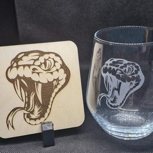 May include: A wooden coaster featuring a laser-etched design of a snake's head. The snake has a white eye and an open mouth with fangs. The coaster is beside a clear glass with a similar snake design etched on the side.