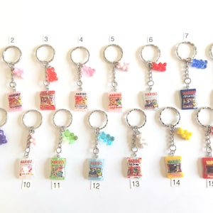 May include: A collection of fifteen keychains, each featuring a miniature Haribo candy bag and a gummy bear charm. The bags display various Haribo product names and colorful graphics. The keychains have silver-colored rings.