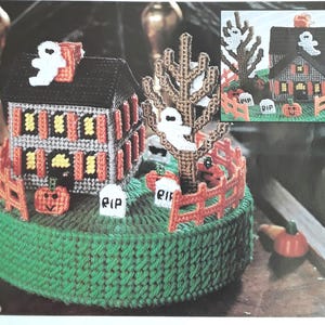 Pode incluir: Uma decoração de Halloween feita à mão com uma casa, uma árvore e uma cerca. A casa tem um telhado preto, persianas laranja e um fantasma branco. Uma árvore com um fantasma e uma cerca circundam a casa. A base é verde com abóboras e lápides.