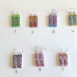May include: Seven pairs of earrings featuring different brands of chewing gum. The earrings are silver and feature a rectangular charm with a colorful image of a chewing gum pack. The brands include Doublemint, Hubba Bubba, Double Bubble, Jolly Rancher, Razzles, Skittles, and Big O.K.