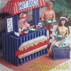 Könnte beinhalten: Ein handgefertigter Miniatur-Imbissstand mit einer rot-weiß gestreiften Markise, auf der "HOT DOGS" steht. Eine Puppe steht hinter der Theke, und zwei weitere Puppen sind in der Nähe. Der Stand ist Teil einer "Boardwalk Picnic"-Szene.