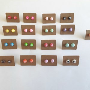 May include: Twenty-four pairs of stud earrings on brown cards. The earrings are small and round, with a white "M" on each. The earrings are in various colors, including red, orange, yellow, green, blue, purple, pink, and brown.