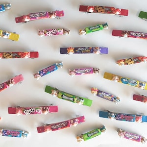 May include: A collection of colorful hair clips featuring various candy bar designs, including Doublemint, Big Hunk, and Crunch. The clips are made of plastic and have a shiny finish.