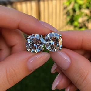May include: A pair of round-cut stud earrings featuring clear, faceted stones set in a four-prong silver-toned metal setting. The stones exhibit high clarity and brilliance, reflecting light with multiple internal facets visible in the circular design.