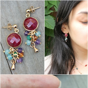 May include: Gold-toned earrings with large, faceted, ruby-red stones as the focal point. Each earring features a cluster of smaller, colorful beads in shades of purple, green, and blue, suspended below the ruby. The earrings are shown in multiple views.
