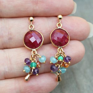 May include: Gold-toned earrings featuring a large, faceted, round, red gemstone at the top. Below, clusters of small, colorful beads in shades of purple, green, blue, and orange dangle from the earrings. The earrings are held up by a person's hand.