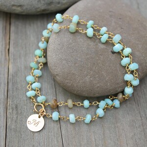 May include: A double-strand necklace with small, pale blue and tan gemstone beads on a gold-coloured chain. A small, round charm with the letter "M" is attached. The jewellery sits on a grey stone and wooden surface.