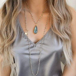 May include: Two layered necklaces with gold chains and grey beads. The top necklace features a teardrop-shaped pendant with a blue-green stone. The bottom necklace is a long strand with a toggle clasp and a small gold disc.